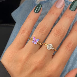 Puede incluir: Dos anillos de oro en una mano. Un anillo presenta un dise&ntilde;o de mariposa con piedras iridiscentes moradas y rosas. El otro anillo tiene una gran piedra transparente de talla ovalada con dos piedras transparentes m&aacute;s peque&ntilde;as. Las u&ntilde;as est&aacute;n pintadas en tonos verdes y rosas.