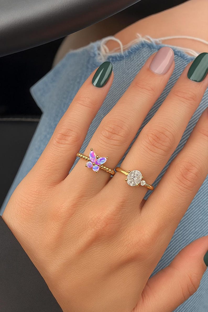 Puede incluir: Dos anillos de oro en una mano. Un anillo presenta un dise&ntilde;o de mariposa con piedras iridiscentes moradas y rosas. El otro anillo tiene una gran piedra transparente de talla ovalada con dos piedras transparentes m&aacute;s peque&ntilde;as. Las u&ntilde;as est&aacute;n pintadas en tonos verdes y rosas.