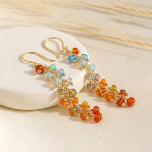 Könnte beinhalten: Ein Paar goldfarbene Hängeohrringe. Jeder Ohrring hat einen goldenen Haken und eine Kaskade farbiger Perlen in Orange-, Rot-, Blau- und Grüntönen. Die Perlen sind gruppiert und erzeugen ein strukturiertes und auffälliges Design.