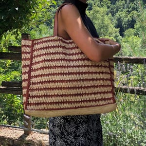 May include: A large, rectangular tote bag with a woven design in alternating stripes of beige and reddish-brown. The bag has brown leather straps and a natural fiber base. The bag is being carried outdoors, with a natural background of trees and a wooden fence.