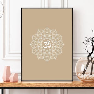 May include: A framed print featuring a white mandala design with the Om symbol on a tan background. The artwork is displayed on a wooden table with decorative vases and a glass vase with branches. The logo "Bird of Juno Creations" is in the upper left corner.