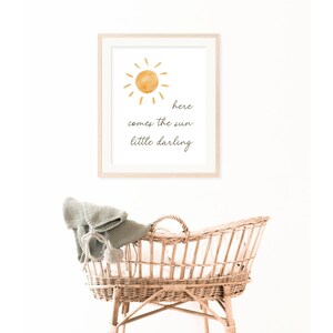May include: A framed print with a watercolor sun illustration and the text "here comes the sun little darling." Below, a woven wicker bassinet is visible, with a gray blanket and pom-pom detail.