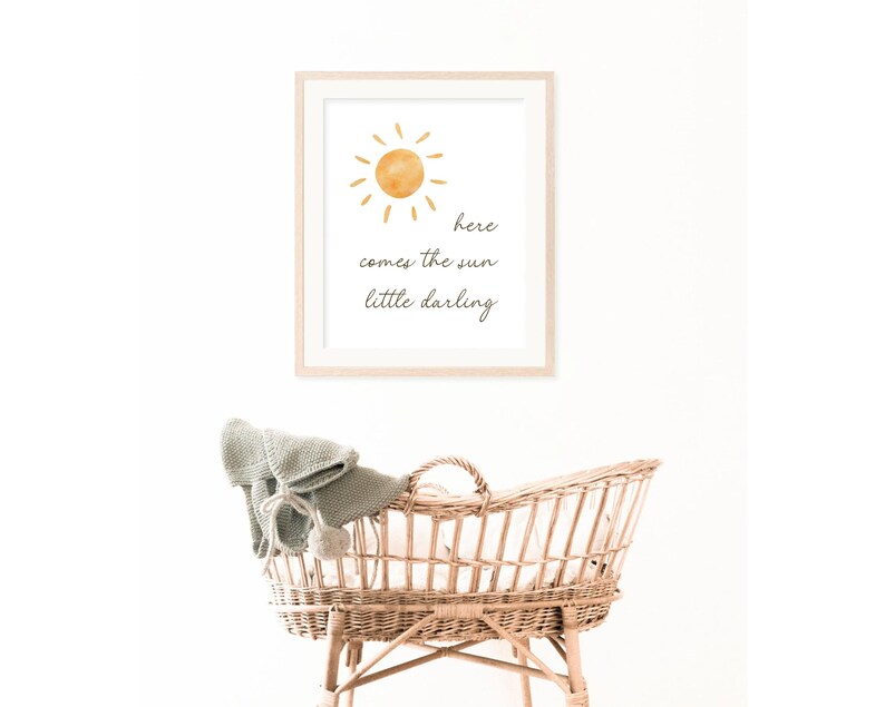 May include: A framed print with a watercolor sun illustration and the text "here comes the sun little darling." Below, a woven wicker bassinet is visible, with a gray blanket and pom-pom detail.