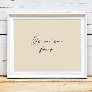 May include: A framed print with the phrase "Je m'en fous" in elegant black script on a beige background. The white frame contrasts with the text and background, creating a minimalist aesthetic. The print is displayed on a wooden surface.