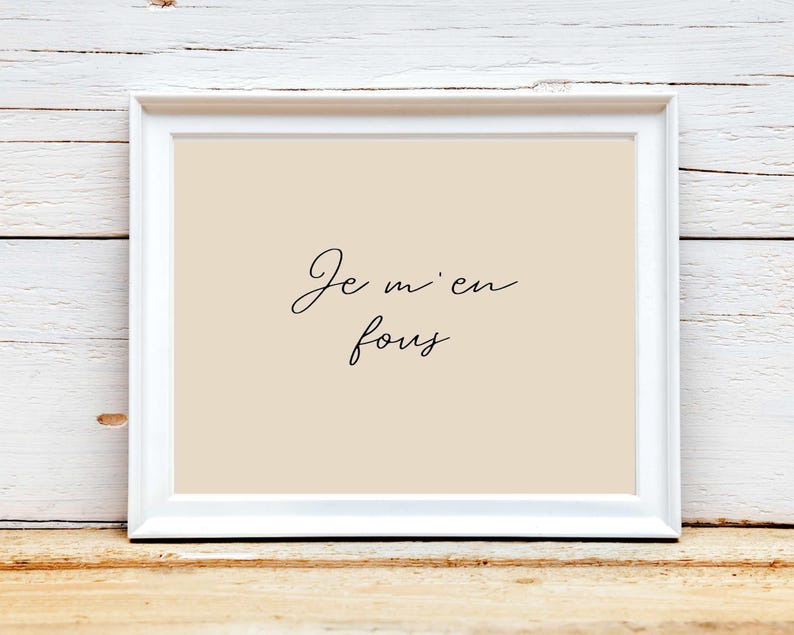 May include: A framed print with the phrase "Je m'en fous" in elegant black script on a beige background. The white frame contrasts with the text and background, creating a minimalist aesthetic. The print is displayed on a wooden surface.