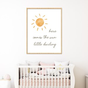 May include: A framed print featuring a watercolor sun illustration in shades of orange and yellow. The text "here comes the sun little darling" is written in a delicate script below the sun. The print is displayed above a white crib.