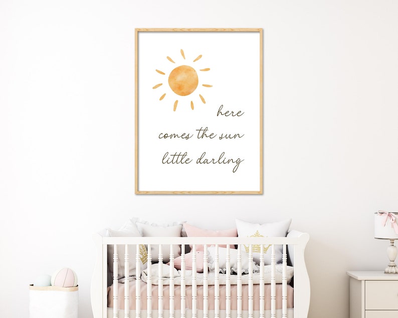 May include: A framed print featuring a watercolor sun illustration in shades of orange and yellow. The text "here comes the sun little darling" is written in a delicate script below the sun. The print is displayed above a white crib.