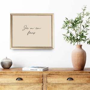 May include: A framed print with the text "Je m'en fous" in elegant script. The artwork has a beige background and a gold frame. A wooden dresser with a decorative object and a vase with greenery are in the foreground.
