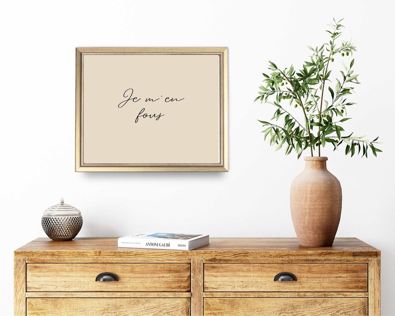 May include: A framed print with the text "Je m'en fous" in elegant script. The artwork has a beige background and a gold frame. A wooden dresser with a decorative object and a vase with greenery are in the foreground.