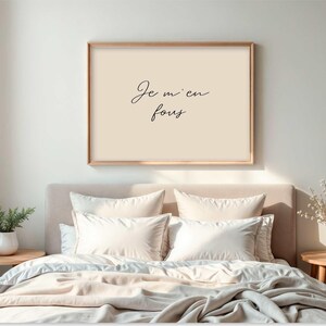 May include: A framed print with the text "Je m'en fous" in a bedroom setting. The print has a light beige background and a wooden frame. The bed is made with white pillows and a light beige comforter. Two small wooden side tables are on either side of the bed.