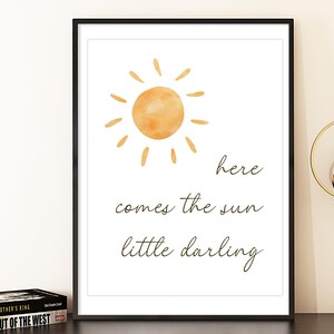 May include: A framed print featuring a watercolor illustration of a sun with rays in shades of orange. The text "here comes the sun little darling" is written below the sun in a handwritten font. The print is in a black frame.