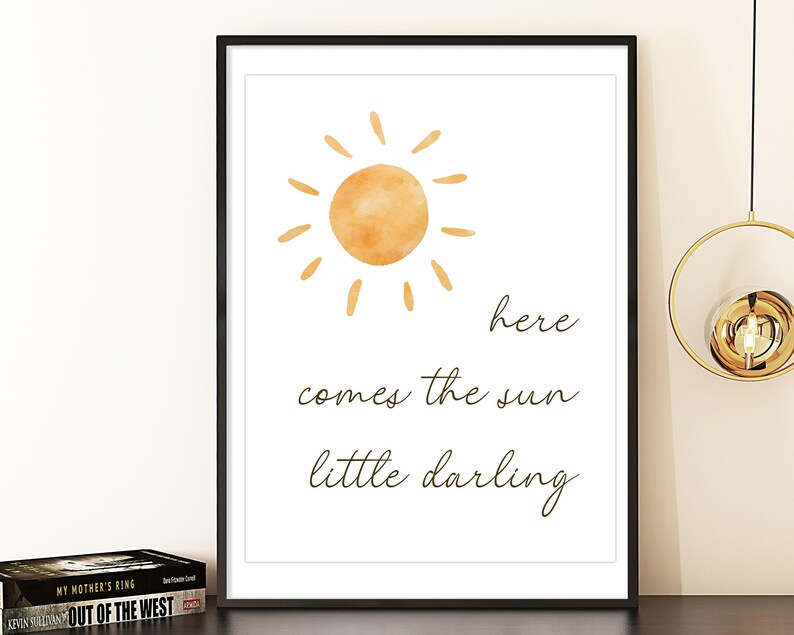 May include: A framed print featuring a watercolor illustration of a sun with rays in shades of orange. The text "here comes the sun little darling" is written below the sun in a handwritten font. The print is in a black frame.