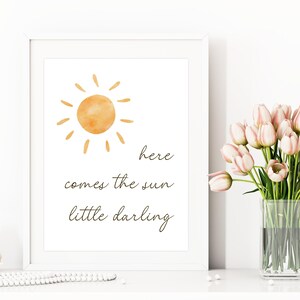 May include: A framed art print with a watercolor sun illustration and the text "here comes the sun little darling." The artwork is in a white frame. A vase of pink tulips sits nearby.
