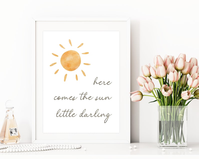 May include: A framed art print with a watercolor sun illustration and the text "here comes the sun little darling." The artwork is in a white frame. A vase of pink tulips sits nearby.