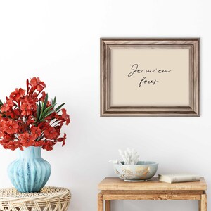 May include: A framed print with the phrase "Je m'en fous" in elegant script. The frame is a warm brown, and the print has a cream-colored background. A blue vase with red flowers sits on a woven table, and a small wooden table holds a bowl and a book.