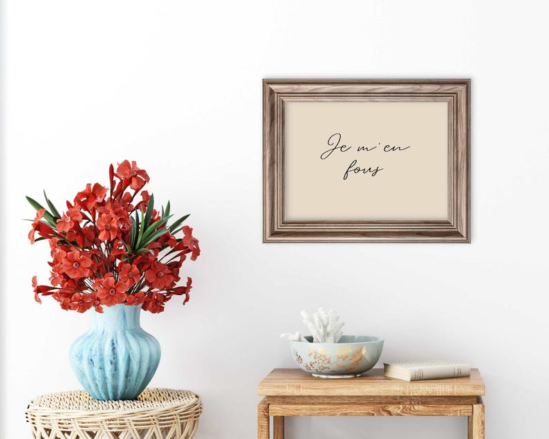 May include: A framed print with the phrase "Je m'en fous" in elegant script. The frame is a warm brown, and the print has a cream-colored background. A blue vase with red flowers sits on a woven table, and a small wooden table holds a bowl and a book.