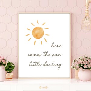 May include: A framed print featuring a watercolor sun illustration in shades of orange. The text "here comes the sun little darling" is written in a delicate script below the sun. The print is displayed on a white surface with pink flowers.
