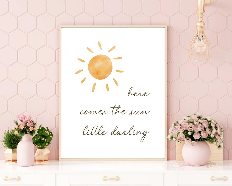 May include: A framed print featuring a watercolor sun illustration in shades of orange. The text "here comes the sun little darling" is written in a delicate script below the sun. The print is displayed on a white surface with pink flowers.
