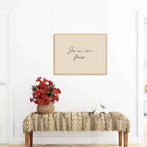 May include: A framed print with the text "Je m'en fous" in black script hangs above a bench. The bench has a woven beige and cream-colored textile. A vase of red flowers sits on the bench, and a white decorative object is also present.