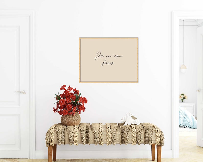 May include: A framed print with the text "Je m'en fous" in black script hangs above a bench. The bench has a woven beige and cream-colored textile. A vase of red flowers sits on the bench, and a white decorative object is also present.