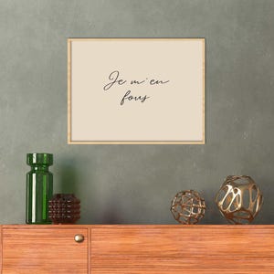 May include: A framed print with the text "Je m'en fous" in cursive script hangs above a wooden cabinet. Decorative items include a green glass bottle, a brown vase, and two gold-colored geometric spheres.