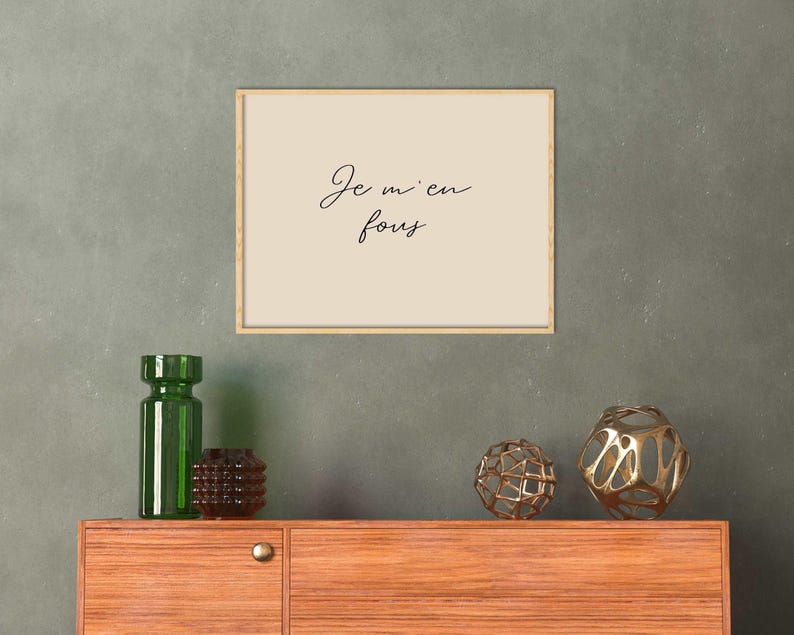May include: A framed print with the text "Je m'en fous" in cursive script hangs above a wooden cabinet. Decorative items include a green glass bottle, a brown vase, and two gold-colored geometric spheres.