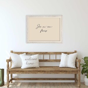 May include: A framed print with the text "Je m'en fous" hangs above a wooden bench with two pillows. The bench has a rustic design, and a potted plant and green boots are on either side.