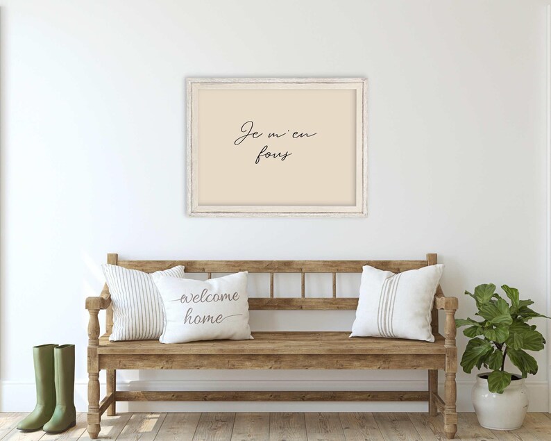 May include: A framed print with the text "Je m'en fous" hangs above a wooden bench with two pillows. The bench has a rustic design, and a potted plant and green boots are on either side.