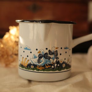 Un pot en émail vintage avec anse décoré d'un motif ludique d'oie/canard, une tasse à mesurer vintage, une cafetière