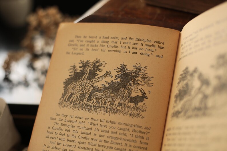 May include: An open book with black and white illustrations of animals. The text reads, "The te been a loud noise, and the Ethioplan called out, "I've caught a thing that I can't see. It smells like Giraffe, but it has no form. "Get on it's head till morning as I am doing," said the Leopard"
