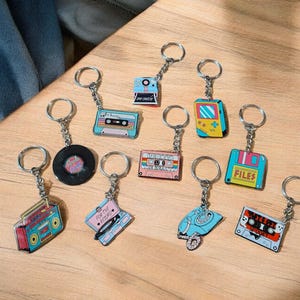 以下が含まれることがあります： カセットテープ、レコード、ラジカセ、電話、携帯ゲーム機などのデザインが施された、レトロをテーマにしたキーホルダーのコレクション。キーホルダーは青、ピンク、黄色など様々な色があり、木製の表面に配置されています。