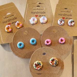 Puede incluir: Una colección de pendientes de botón hechos a mano con forma de donut en varios colores, incluyendo rosa, azul y marrón, expuestos en tarjetas de pendientes de cartón marrón. Algunos pendientes tienen detalles de chispas y glaseado. Las tarjetas dicen "Handmade with love."