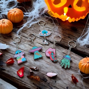 May include: A collection of novelty keychains with alien-themed designs. The keychains feature colorful UFOs, aliens, and other quirky elements. The background includes pumpkins, spiderwebs, and fall foliage, creating a Halloween or spooky theme.