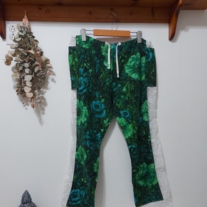 May include: Green floral print pants with white lace trim hanging on a wooden hanger. The pants feature a drawstring waist and a vibrant pattern of green and blue flowers. The lace trim runs along the outer seams of the pants.