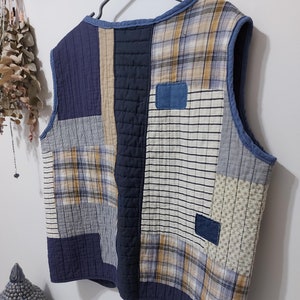 May include: A quilted vest with a patchwork design. The vest features a variety of fabrics in shades of blue, beige, and white, including plaid, striped, and solid patterns. The vest is sleeveless and has a blue trim.
