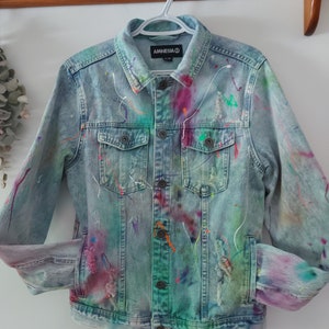 May include: A light blue denim jacket with paint splatters in various colors, including pink, purple, green, and orange. The jacket has a distressed look with rips and tears. The label on the jacket reads "Amnesia".