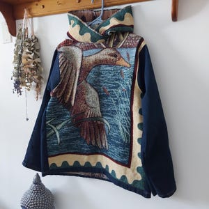 May include: A blue denim jacket with a tapestry panel featuring a duck flying over water. The tapestry is framed with a yellow, green, and red border.