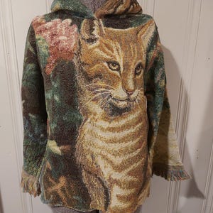 May include: A green, brown, and beige tapestry hoodie with a cat design. The cat is looking directly at the viewer. The hoodie has a hood and long sleeves.