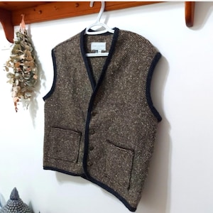 Puede incluir: Un chaleco de tweed marrón con ribete azul oscuro y dos bolsillos delanteros. El chaleco tiene cuello en V y está abotonado. El chaleco está colgado en una pared blanca.