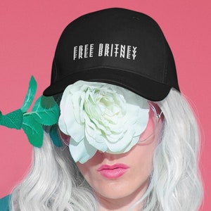 May include: Black baseball cap with the text "FREE BRITNEY" in white. The cap is worn by a person with white hair, a white flower, and a pink background. The flower covers the person's eyes.
