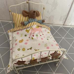 Puede incluir: Una cama de metal blanco en miniatura con un muñeco de ratón de peluche. La cama tiene una almohada a rayas amarillas y blancas y una manta estampada con un unicornio rosa, flores y pájaros. Estrellas doradas decorativas adornan el pie de la cama.