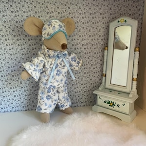 Può includere: Un peluche a forma di topo che indossa un pigiama a fiori blu e bianchi con mascherina coordinata e un nastro azzurro. Accanto al topo si trova una piccola toeletta in legno azzurro con un cassetto decorato da un motivo floreale verde.