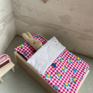 Op de afbeelding: Een miniatuurbed met een roze en witte ruit, versierd met uilen- en hartmotieven. Een klein beige konijn rust op het kussen. Het bed is gemaakt van hout en heeft een wit gewatteerd deken. Een gouden glitterster hangt erboven.