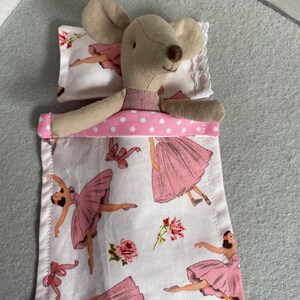 Puede incluir: Una muñeca de ratón beige metida en una cama en miniatura. La ropa de cama presenta un fondo blanco con bailarinas rosas, lazos y rosas. La cama tiene un ribete de lunares rosas y una almohada con ribete de encaje blanco.