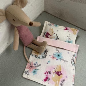 Puede incluir: Un pequeño ratón de peluche con una camisa rosa se sienta junto a un saco de dormir con el tema de Winnie the Pooh. El saco de dormir está hecho de tela blanca con un patrón floral y presenta a Winnie the Pooh y Piglet. El saco de dormir tiene una almohada rosa con Winnie the Pooh en ella.