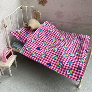 Puede incluir: Una cama de metal blanco en miniatura con una manta a cuadros rosa y blanca con diseños de búhos y corazones. Un peluche beige descansa sobre la almohada. Una pequeña radio rosa está sobre una mesita.