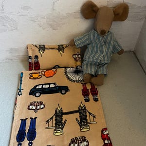 Puede incluir: Un juego de cama de muñeca en miniatura con una almohada y una manta beiges con ilustraciones de Londres, como un taxi negro, el Tower Bridge y un autobús rojo de dos pisos. Una pequeña muñeca ratón con pijama a rayas azules y blancas está al lado.