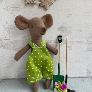 Könnte beinhalten: Eine braune Mauspuppe mit grünen Overalls mit weißen Punkten. Die Puppe hält Miniatur-Gartenwerkzeuge und einen kleinen weißen Topf mit rosa und lila Blumen. Eine goldene Glitzer-Sternengirlande hängt darüber.