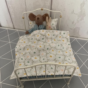 Könnte beinhalten: Ein Miniatur-Metallbett in Weiß mit einer Bettdecke und einem Kissen mit Gänseblümchenmuster. Eine Stoffmaus in blau gestreiftem Pyjama und ein Stoffkaninchen liegen im Bett. Der Bettrahmen hat goldfarbene Details.