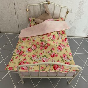 Puede incluir: Una cama de muñeca de metal blanco con un juego de cama estampado floral amarillo y rosa. Un juguete de peluche está acostado en la cama.